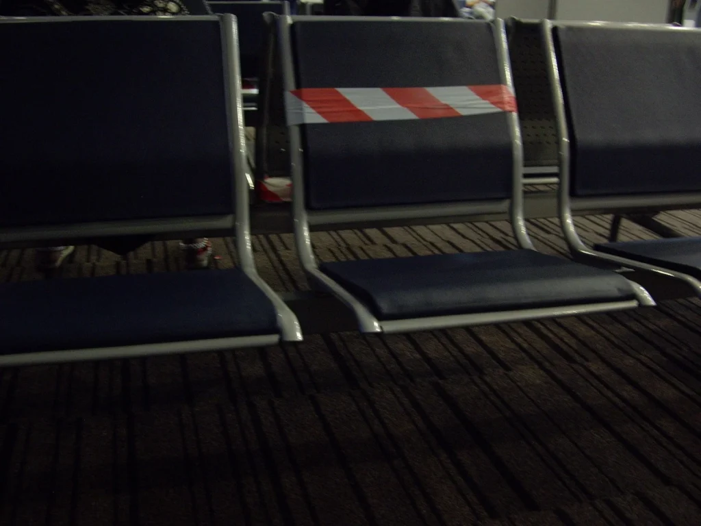 Three airport waiting seats, with a do-not-enter ribbon on the middle one — representing passengers affected by Turkey entry bans waiting for legal resolution.